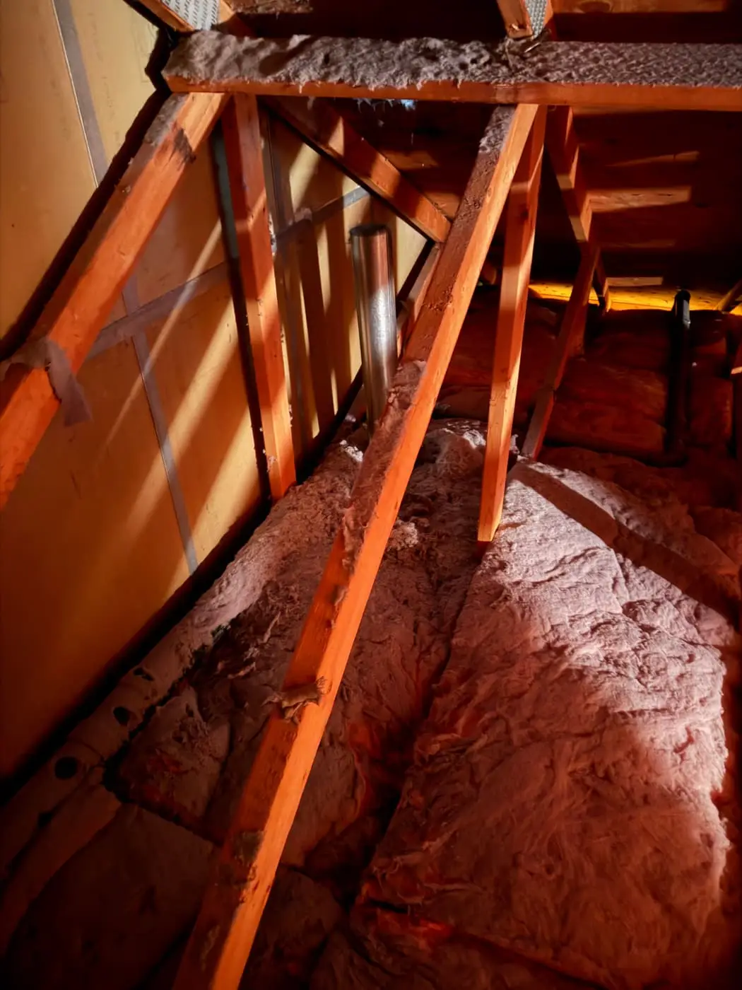 Un tubo de ventilación que sube entre vigas de madera, sobre aislamiento rosado, en un ático con poca luz.
