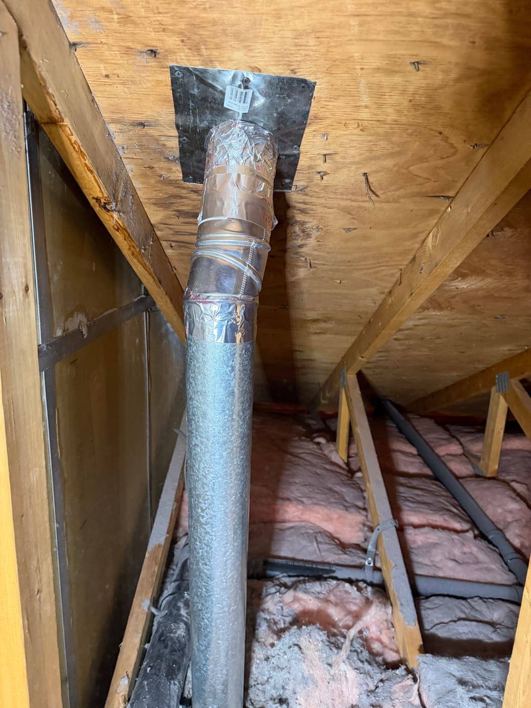 Un tubo de ventilación metálico rígido con cinta de aluminio en las uniones, que sube entre el aislamiento hasta la cubierta del techo de un ático.