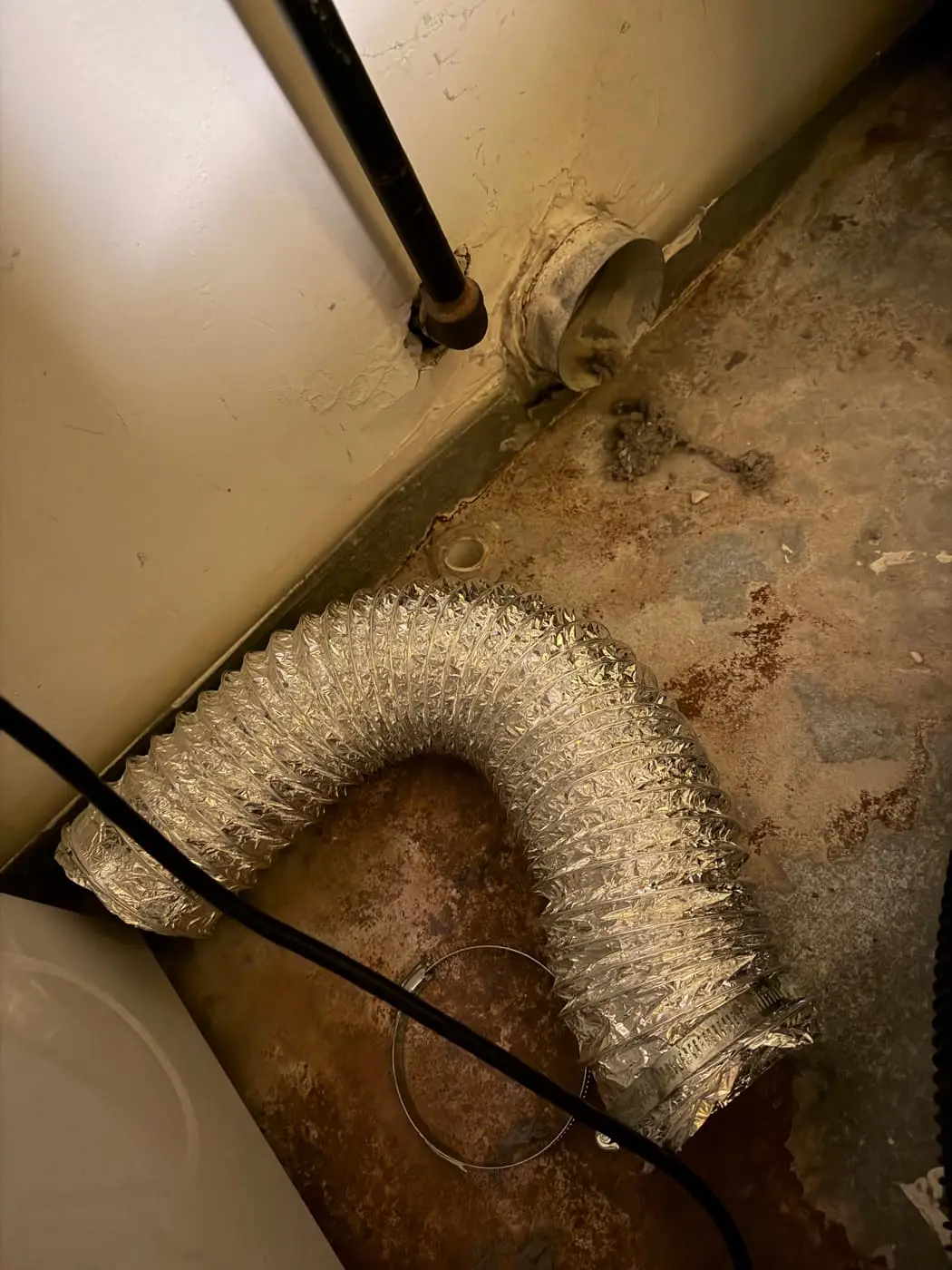 Una manguera flexible de aluminio para secadora en el piso, desconectada del cuello de ventilación de la pared, con pelusa alrededor.