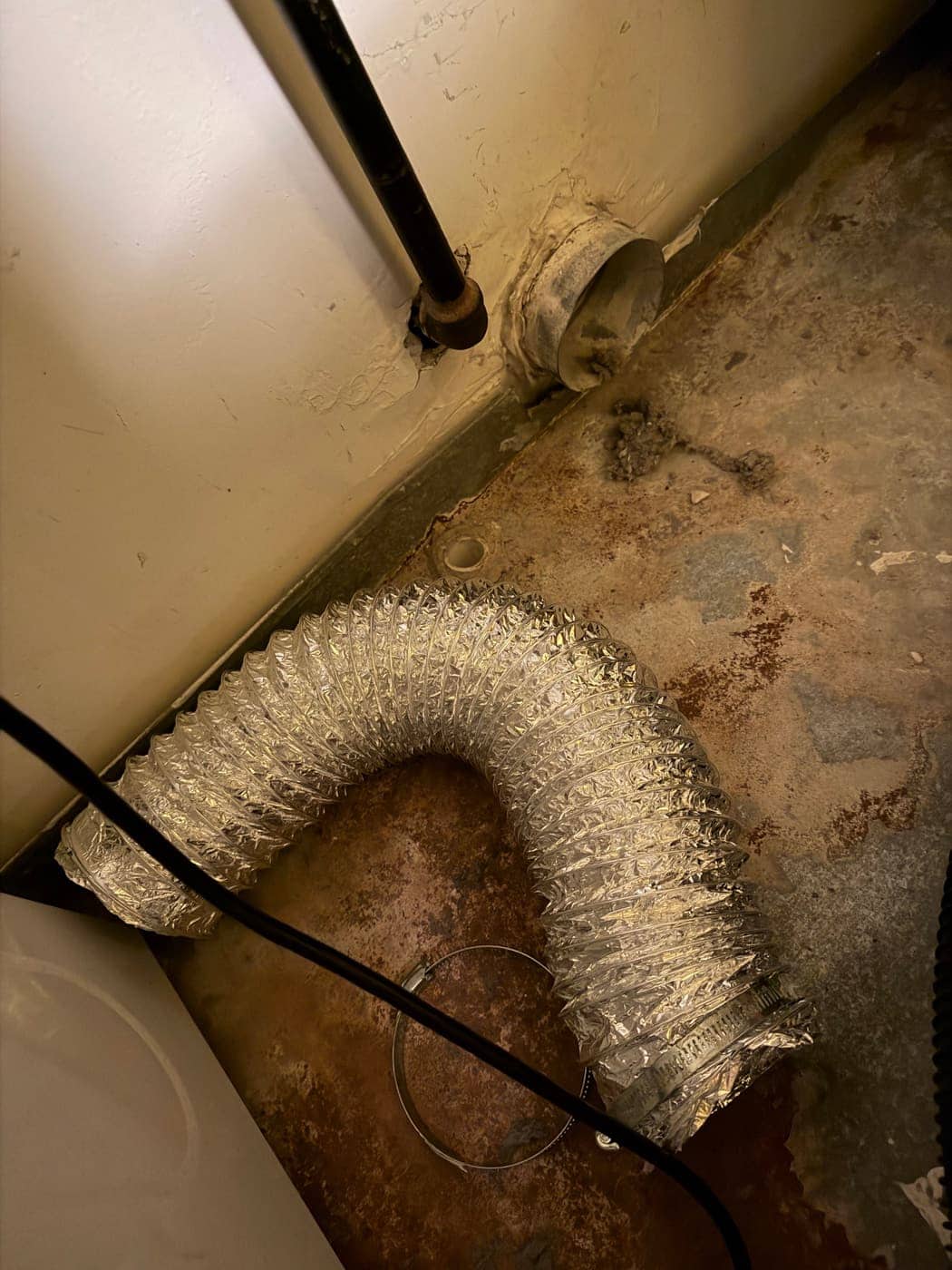 Una manguera flexible de aluminio para secadora en el piso, desconectada del cuello de ventilación de la pared, con pelusa alrededor.