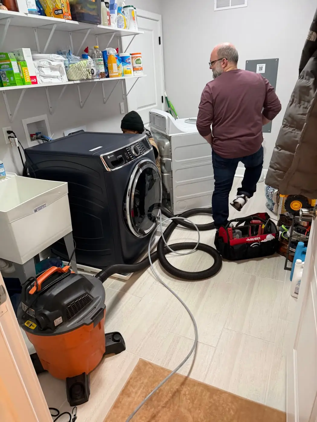 Un cuarto de lavado con la secadora separada de la pared y una aspiradora con mangueras en el piso. Junto a las máquinas hay dos personas.