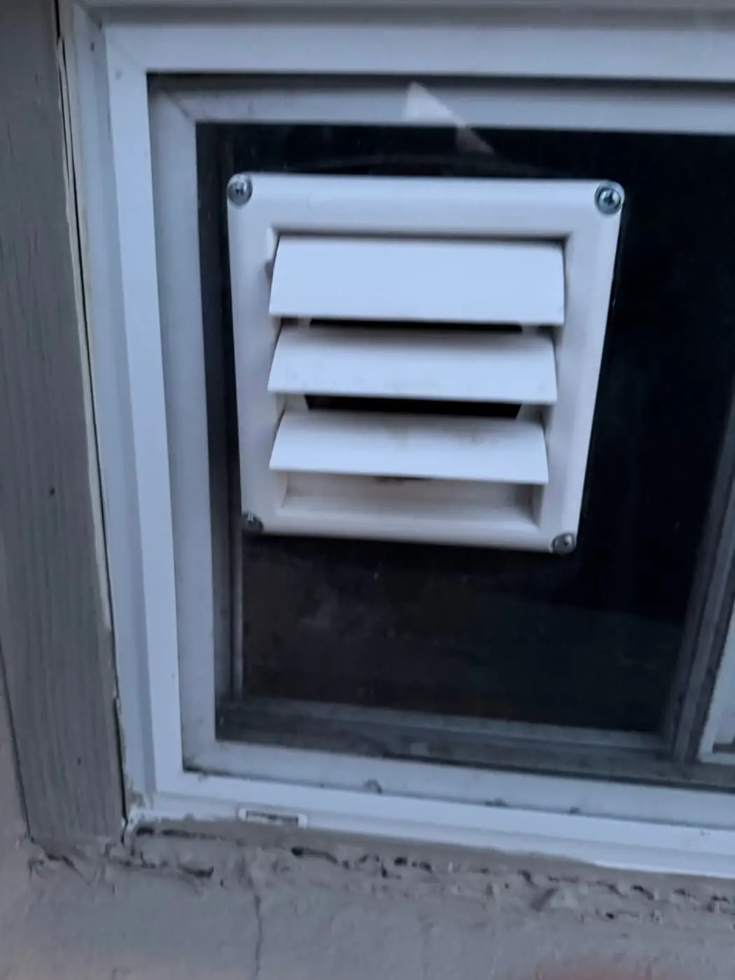 Una salida de ventilación blanca con persianas, colocada en un panel que ocupa la ventana de un sótano.