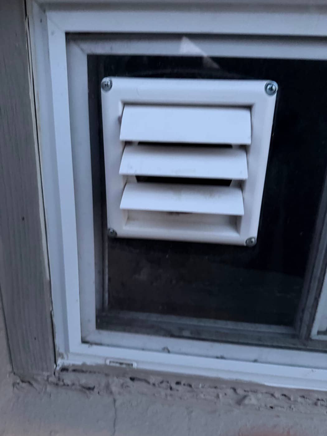 Una salida de ventilación blanca con persianas, colocada en un panel que ocupa la ventana de un sótano.